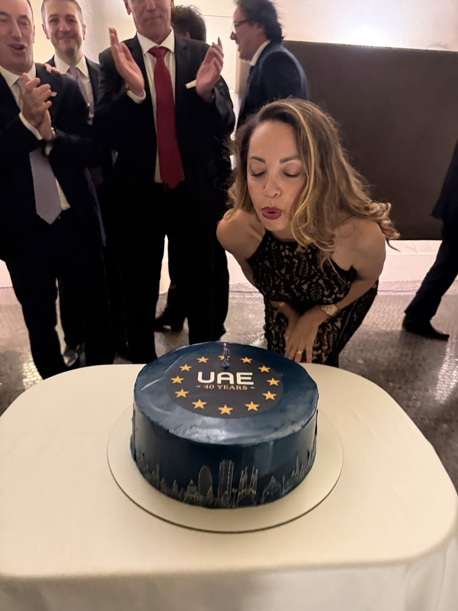 Woman blowing out candles on a blue 40th-anniversary cake as colleagues applaud