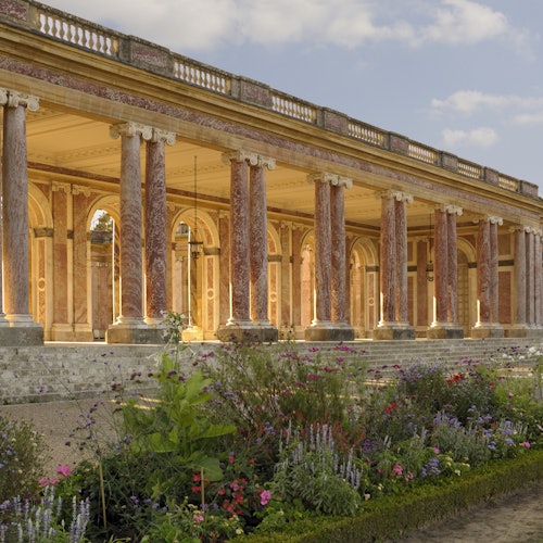 Una gran columnata con columnas y arcos de mármol rosa, enmarcada por un jardín de flores meticulosamente cuidado con plantas vibrantes.