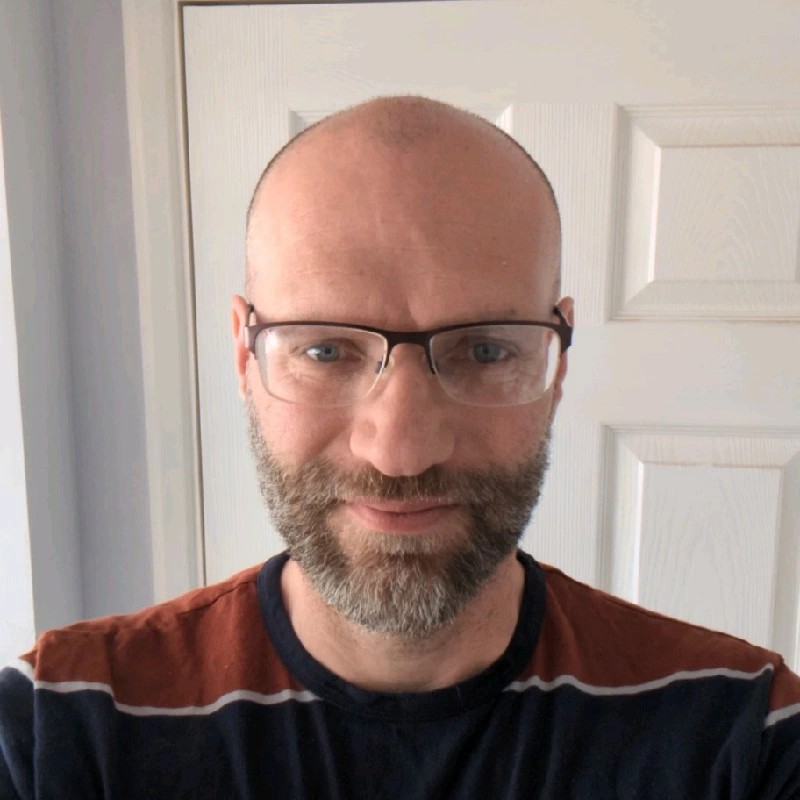 Headshot of a person with glasses and a short beard, wearing a dark top with red detail, standing indoors against a white door.