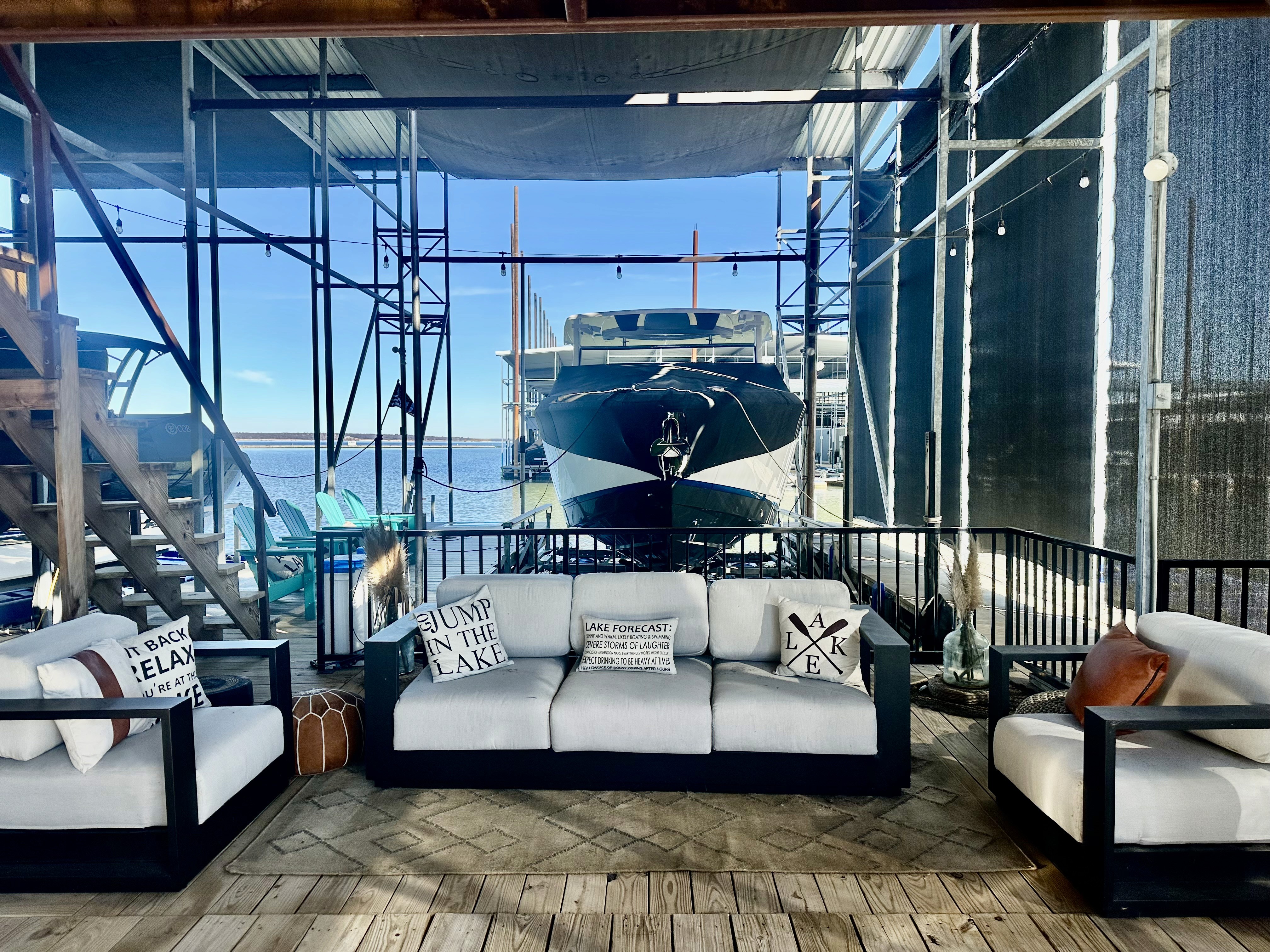 A sleek motorboat is docked in a sunlit marina, surrounded by stylish outdoor seating featuring white cushioned sofas and decorative lake-themed pillows, creating a serene waterfront setting.