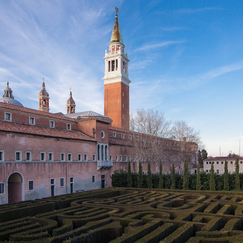 Un labirinto di siepi formale davanti a un grande edificio storico con un alto campanile sotto un cielo azzurro.