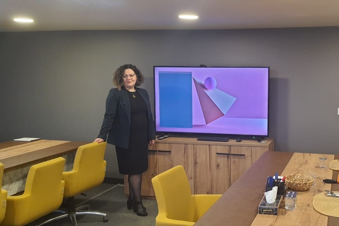 A young person with curly hair smiles at the camera while sitting in front of a laptop against a dark background.