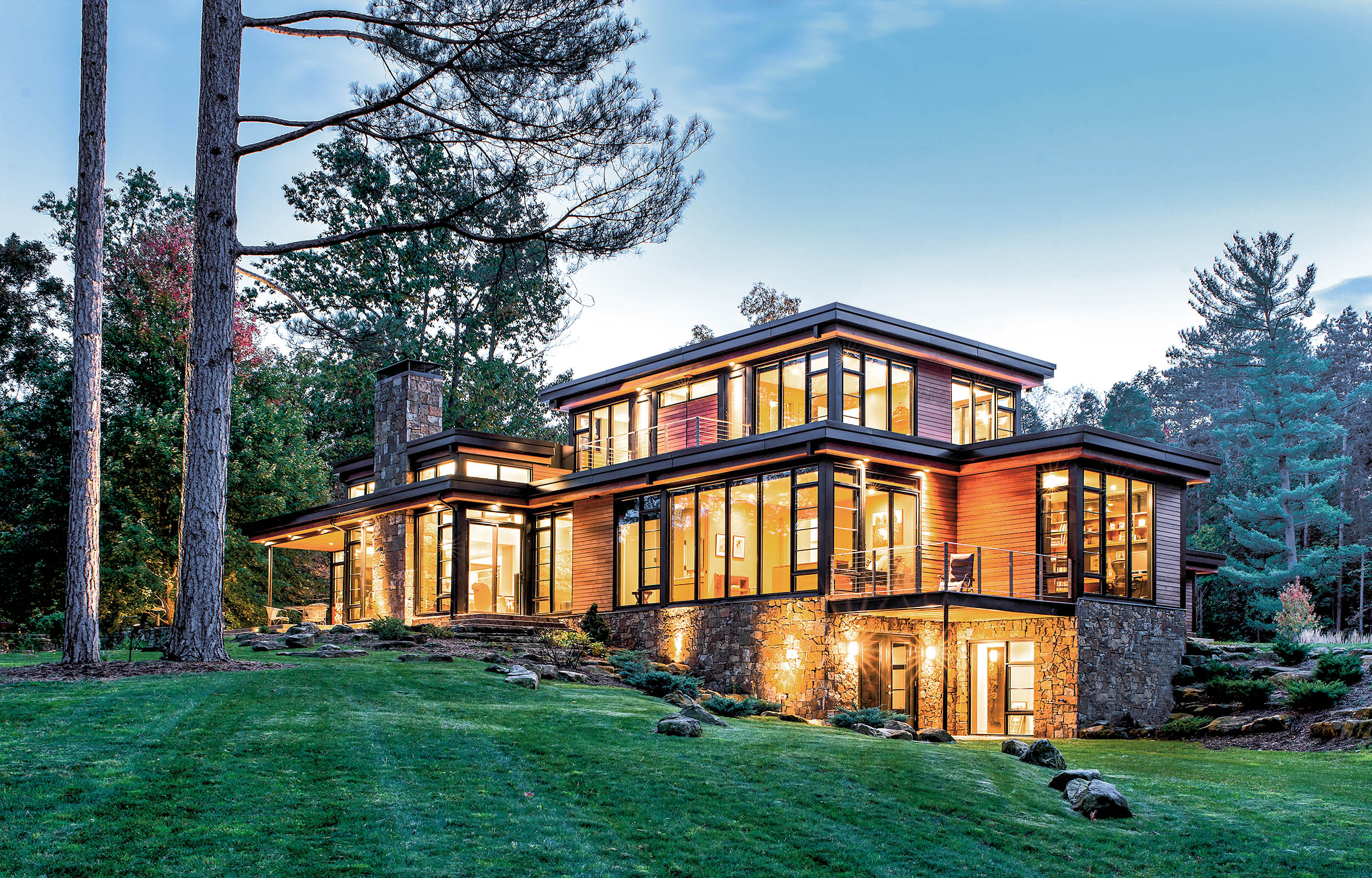 This image features a luxurious, multi-level contemporary home at dusk, designed with a focus on natural materials and transparency. The architecture utilizes a combination of warm wood horizontal siding, natural stone masonry, and expansive black-framed windows.