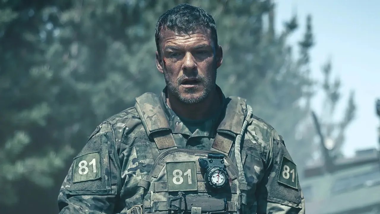 A rugged soldier wearing camouflage fatigues with the number 81 on his gear stands amidst a smoky battlefield, surrounded by trees and military vehicles in the background.