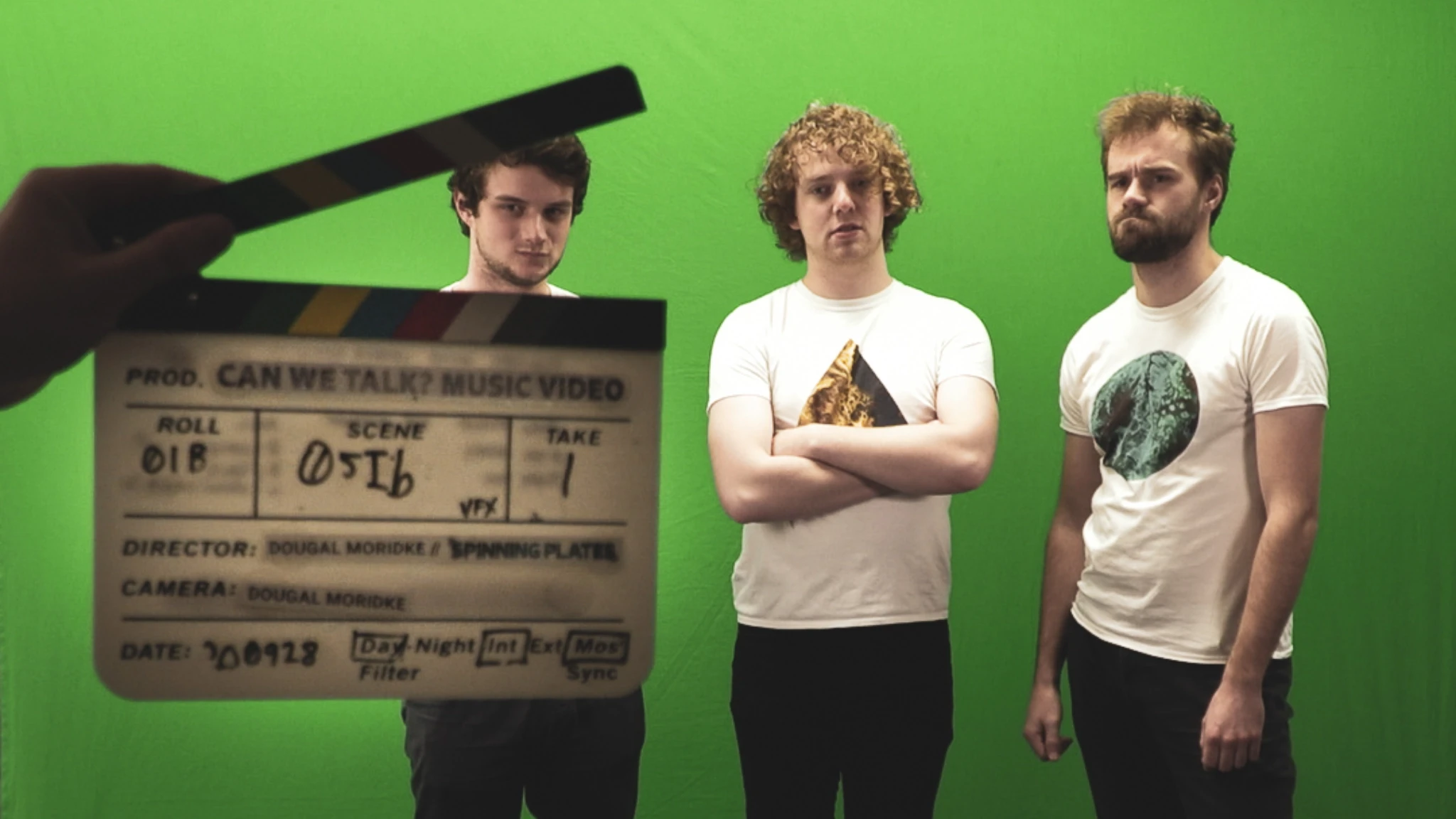 Three people stand against a green screen, a clapperboard primed to snap.