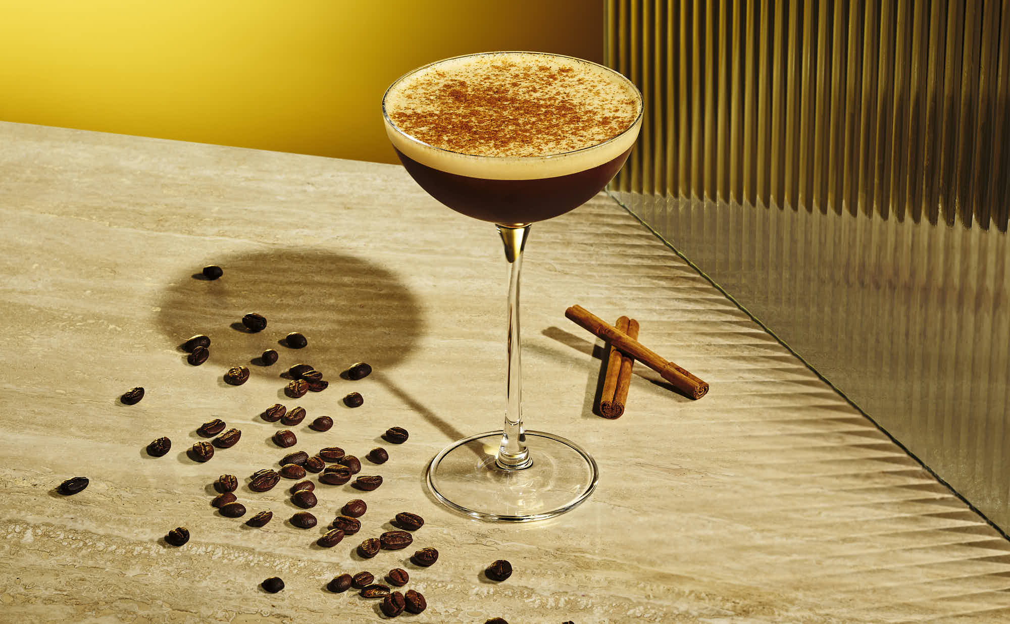 A cocktail glass filled with a brown drink, surrounded by coffee beans and cinnamon sticks on a wooden surface.