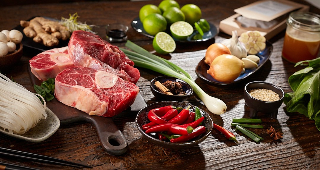 A rustic wooden table is adorned with fresh ingredients for a Vietnamese pho, including raw beef shanks, rice noodles, red chili peppers, limes, ginger, garlic, onions, scallions, mushrooms, hoisin sauce, and star anise, evoking a rich culinary ambiance.