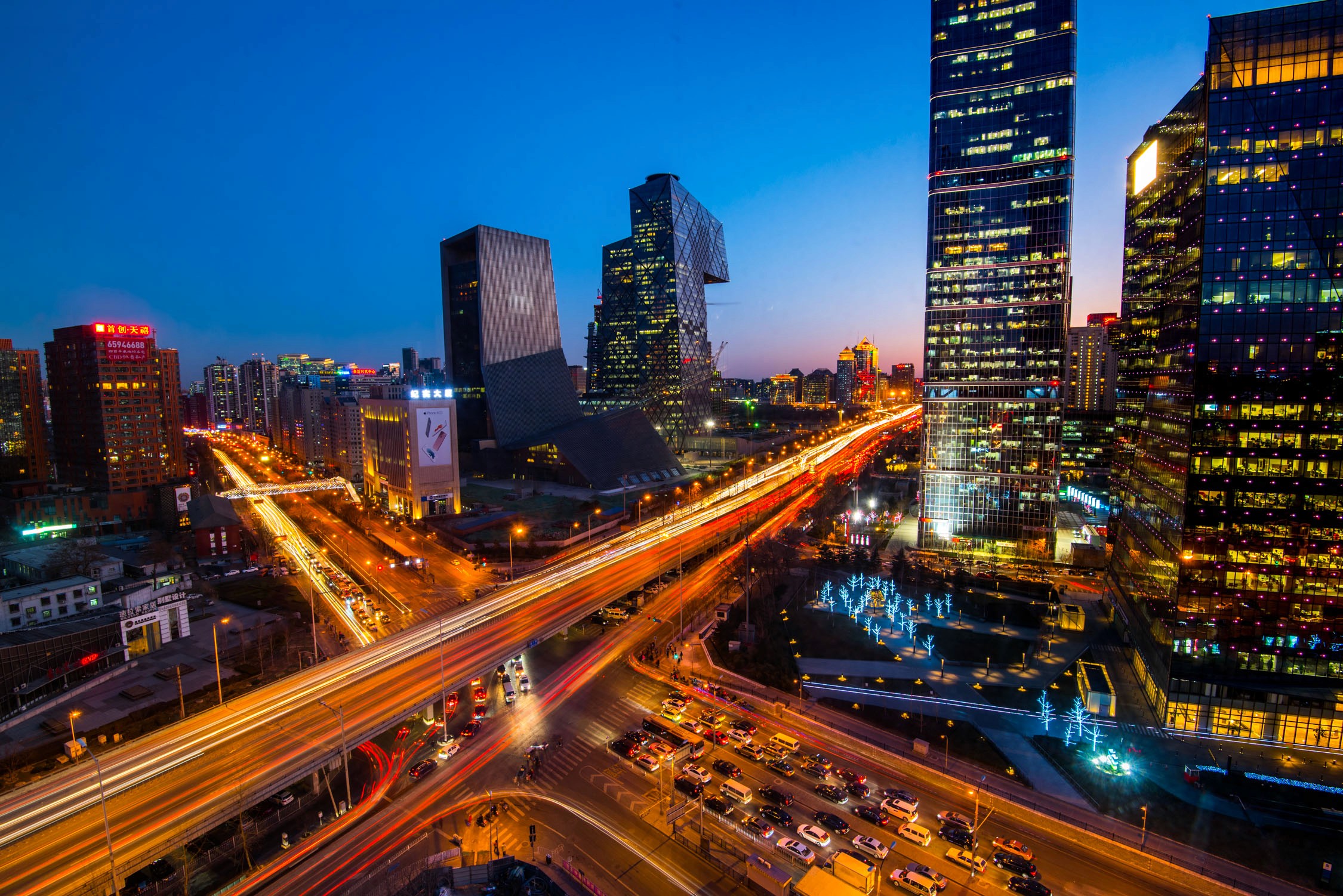 A high-angle night view of a busy multi-lane highway with light trails from cars passing through a city skyline.