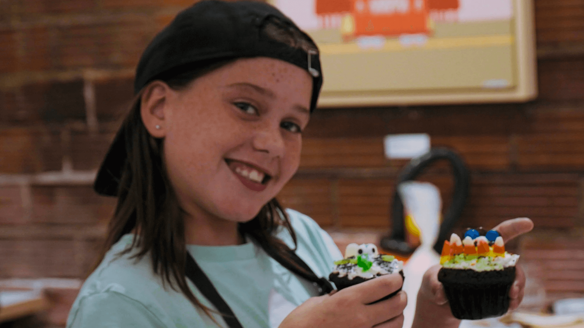 Smiling child wearing a backward baseball cap holding two decorated cupcakes with colorful candy toppings indoors.
