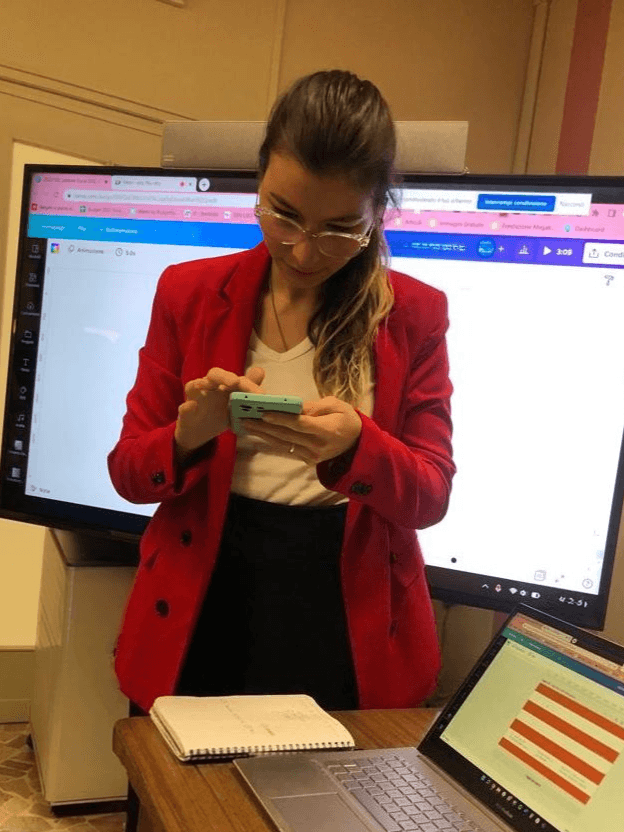 Silvia Vazzana in piedi guarda il cellulare mentre davanti a sé e dietro presenta una presentazione