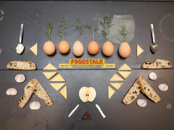 A dark gray surface displays an arrangement of ingredients and utensils for Foodtalk. Five brown eggs, each with a small green sprout, are positioned in a row. Below them, triangular slices of cheese are arranged in two rows. To the right, slices of bread are placed side by side, and to the left, thinly sliced red onion and thin white onion pieces are arranged. Scattered around the arrangement are small pieces of black pepper and salt. A yellow banner in the center reads “FOODSTALK” with “New York” and “Est. 2020” printed below it. Two spoons are placed on either side of the arrangement.