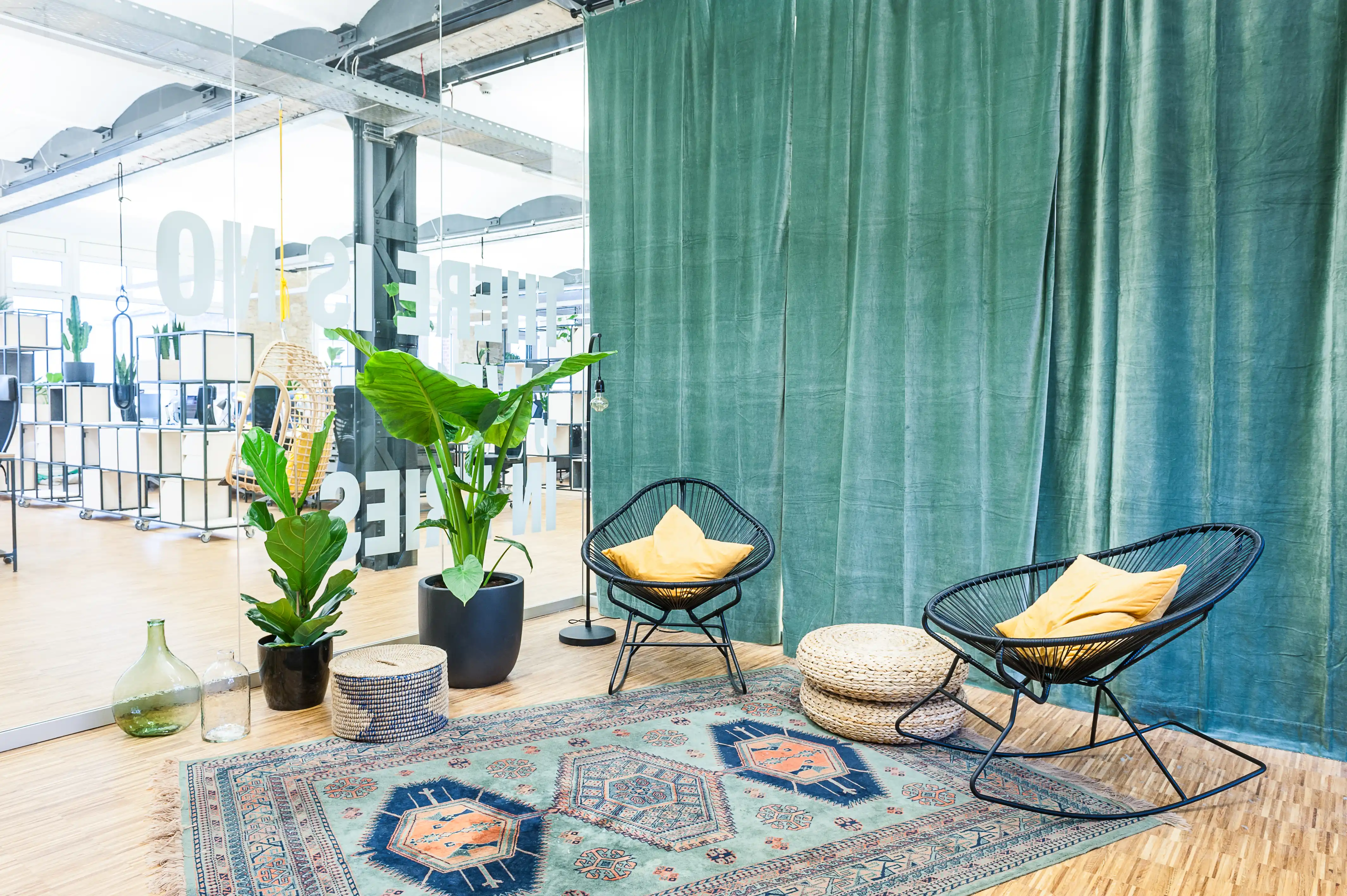 Lounge corner at INTERMATE x TRUEMATES with a floor-length green velvet curtain, two black string chairs with yellow cushions, woven poufs, potted plants and an oriental-style rug.