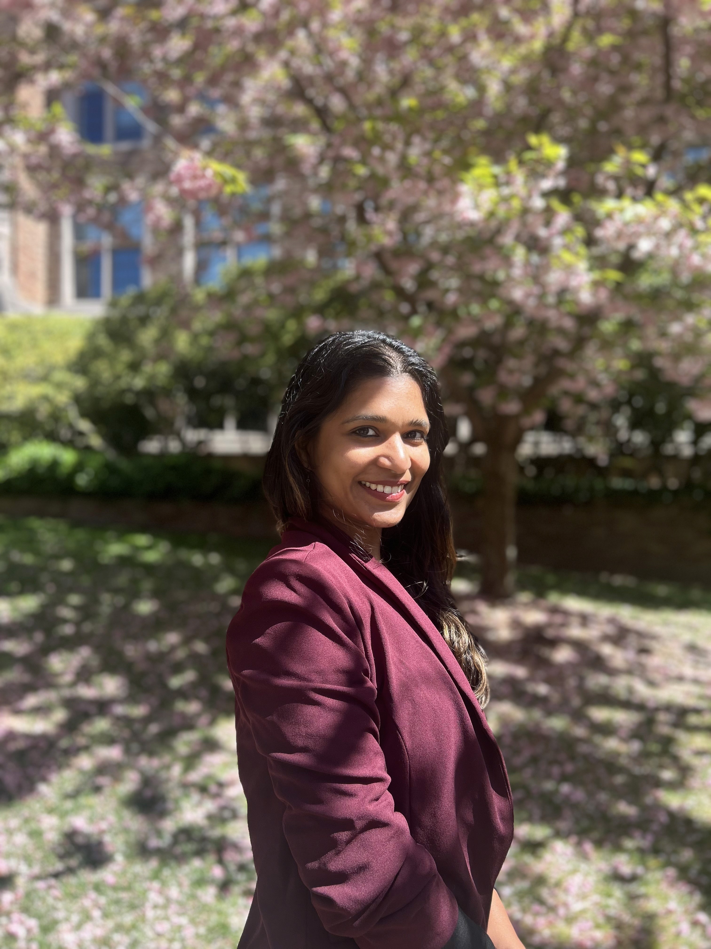 An indian woman with long black hair wearing a burgundy suit jacket. She stands in front of cherry blossom trees at the University of Washington