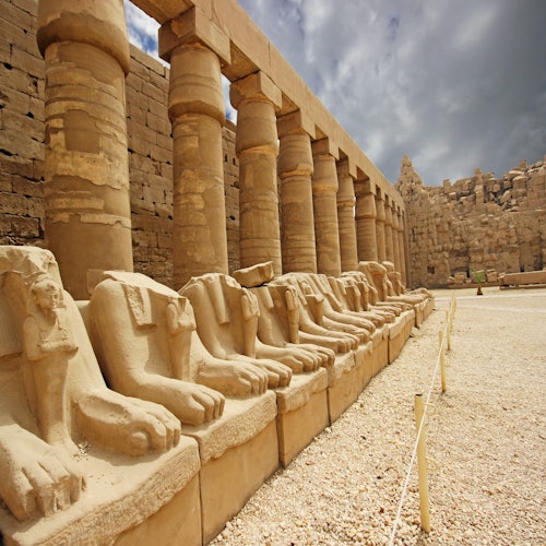 Row of ancient stone sphinx statues with lion bodies and ram heads beside large columns in a historic ruin under a cloudy sky.