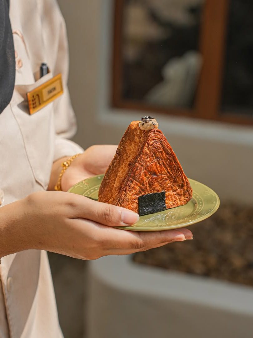A person holding a decorative plate with a pyramid-shaped cake that resembles a traditional house, featuring a black base and some decorative toppings.