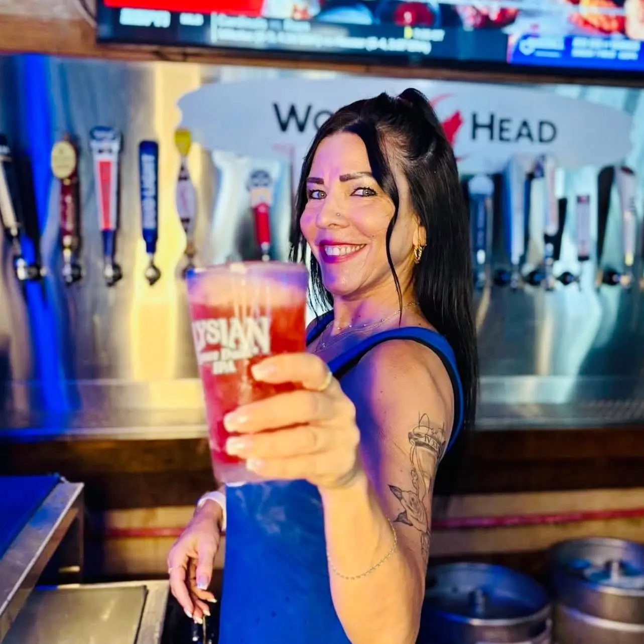 In a lively bar setting in Lakewood Ranch, FL, a smiling woman in a blue tank top, visible tattoos on her arm, holds out a large frothy beer towards the camera, with a row of colorful beer taps behind her.