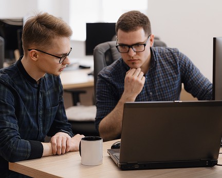 Two people in a modern office setting, focused intently on a computer screen, with one person holding their chin thoughtfully and a coffee mug placed on the desk beside the laptop.