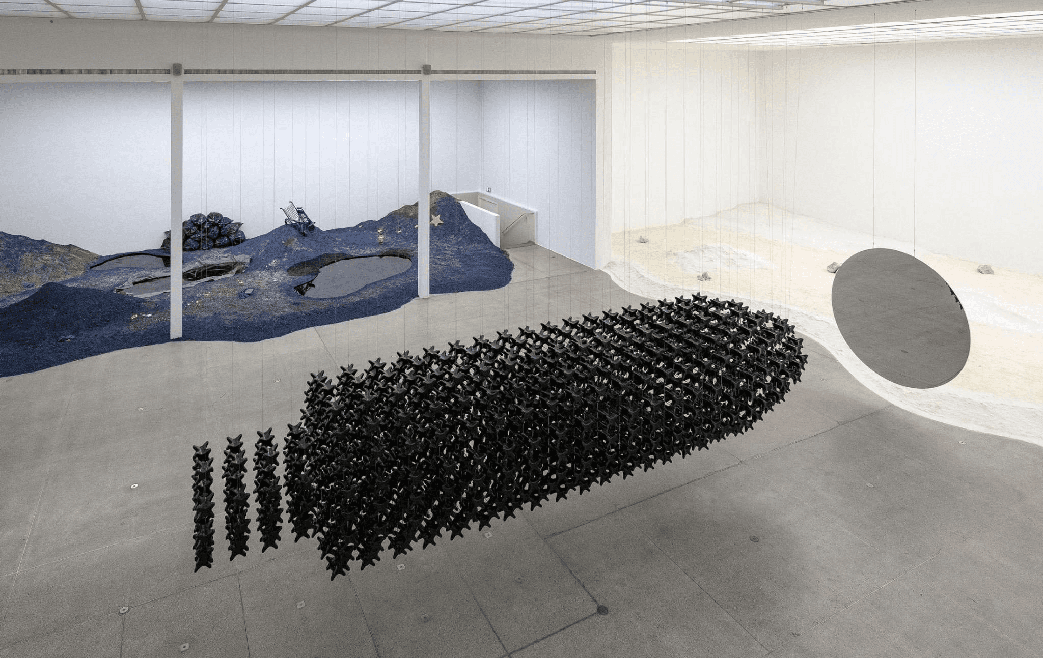 Installation view of Ndidi Dike’s Rare Earth Rare Justice at Secession, Vienna, showing a large suspended formation of black autopsy neck rests, a circular mirror and cobalt-blue and pale mineral-like terrains.