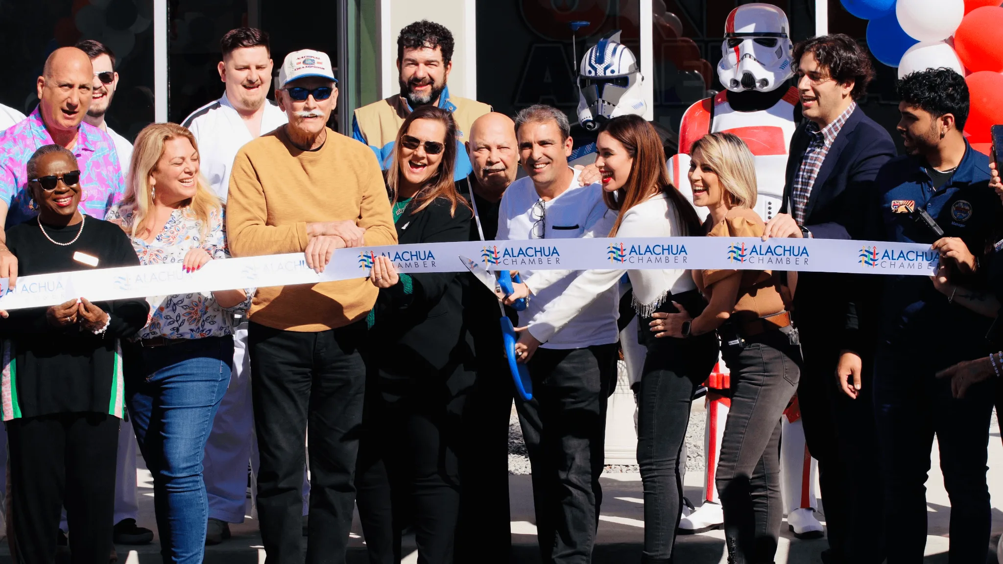 A diverse group of people participate in an Alachua Chamber ribbon-cutting ceremony at a business opening, holding a white banner with the chamber's logo while smiling in bright sunlight.