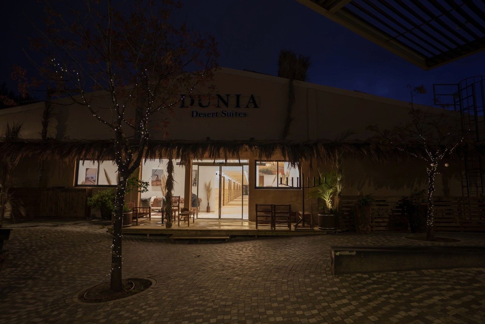 Outdoor lounge under a wooden pergola, draped linen curtains, beige cushions, ocean backdrop, palm shadows dancing on the walls, serene luxury.
