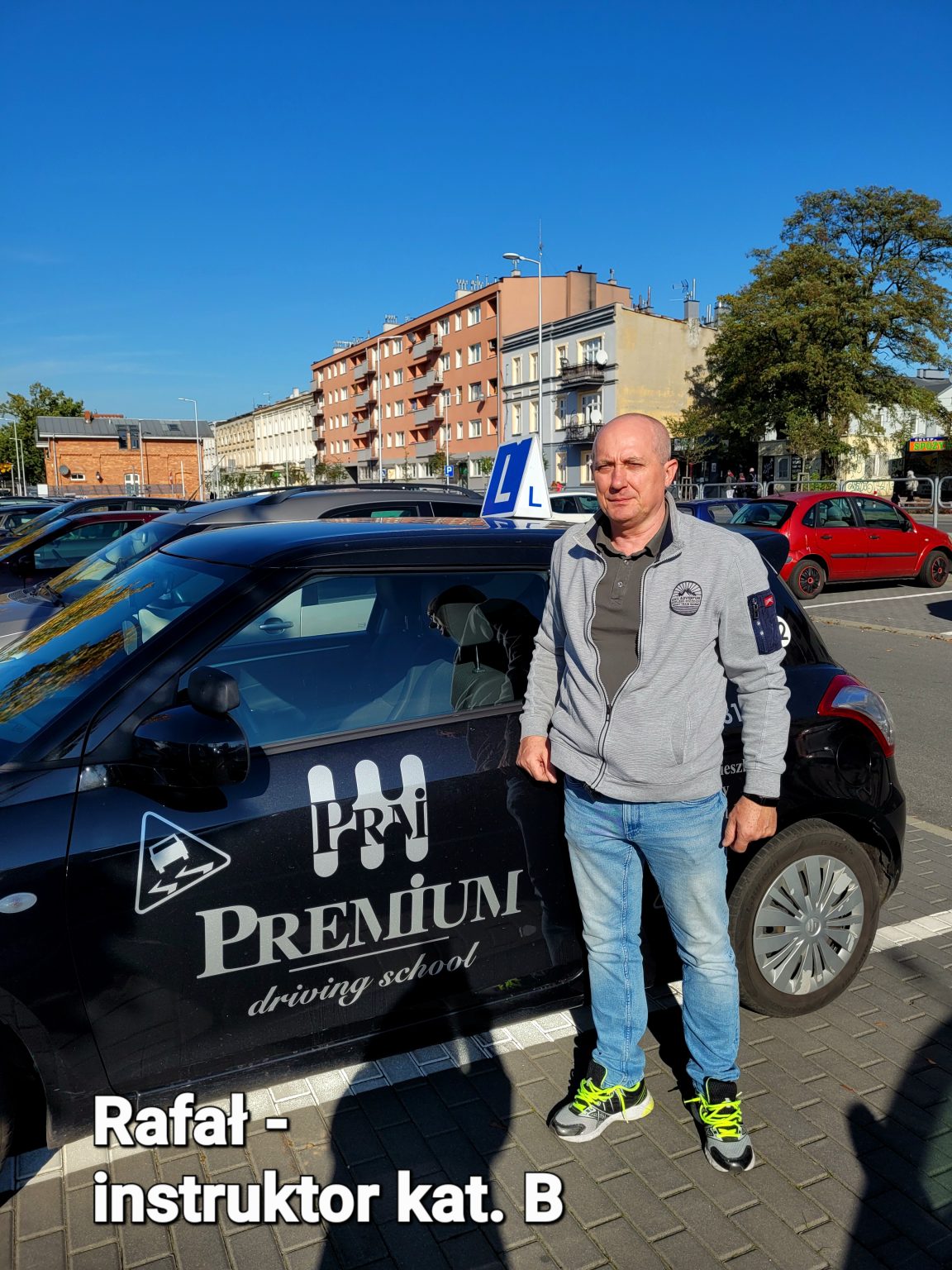 Rafał – Instruktor stojący obok czarnego auta szkoleniowego Premium na tle osiedlowej ulicy.