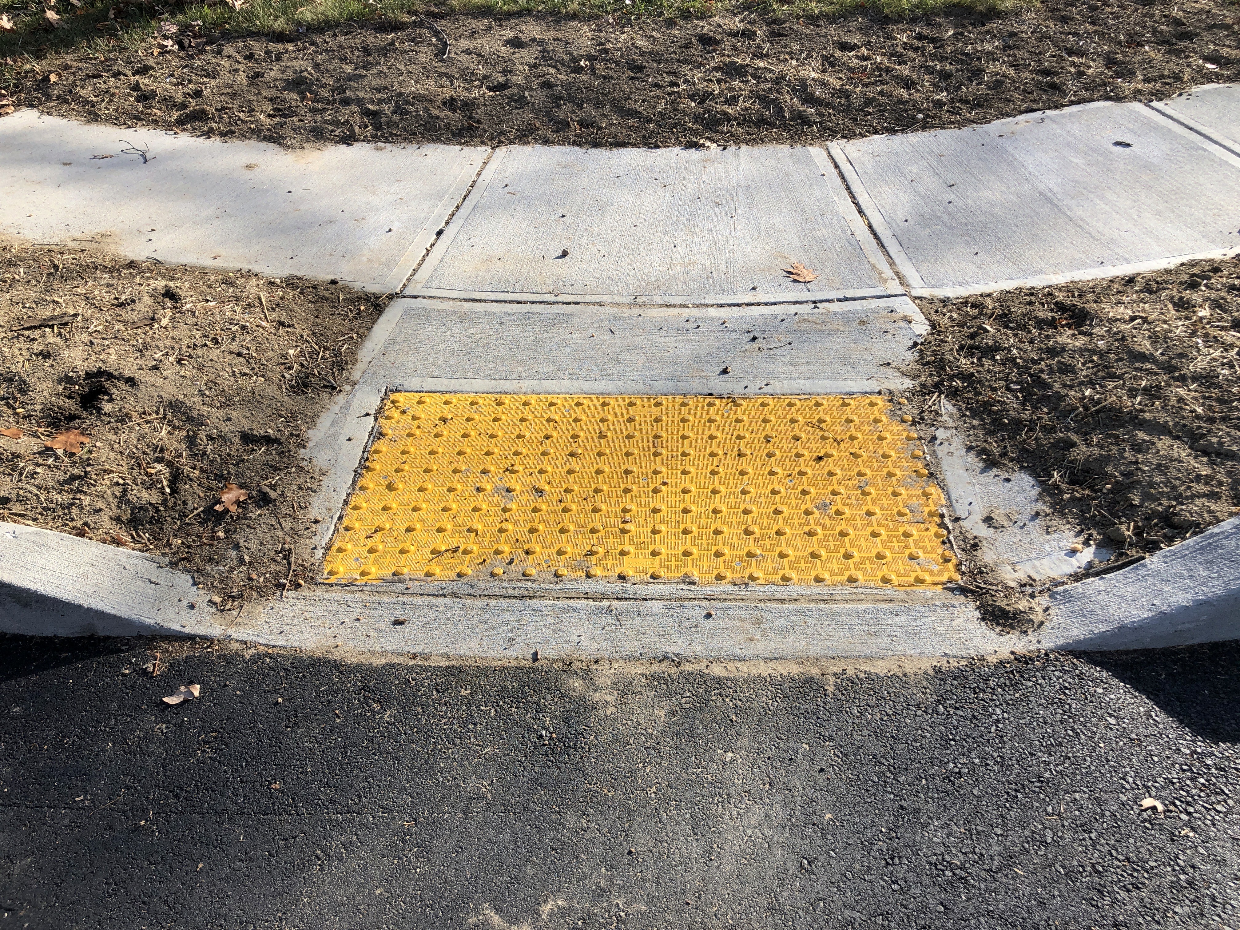 A high-angle view of a newly constructed concrete pedestrian curb cut at a street corner. The ramp leads from a curved sidewalk down to a dark asphalt road. A bright yellow tactile paving tile with a raised dome pattern is embedded in the centre of the ramp for accessibility.