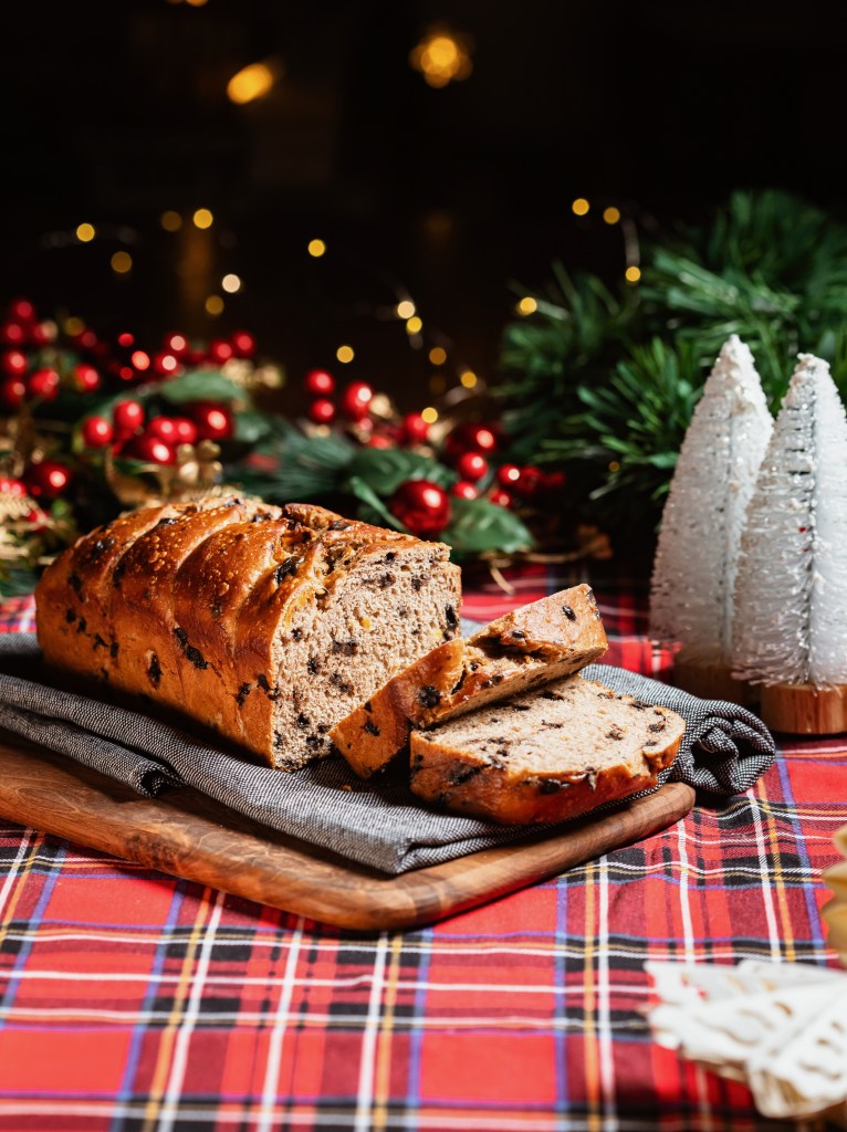 A loaf of festive fruit bread sliced on a wooden board, with Christmas decorations in the background, including small white trees and red berries, set against a plaid tablecloth.