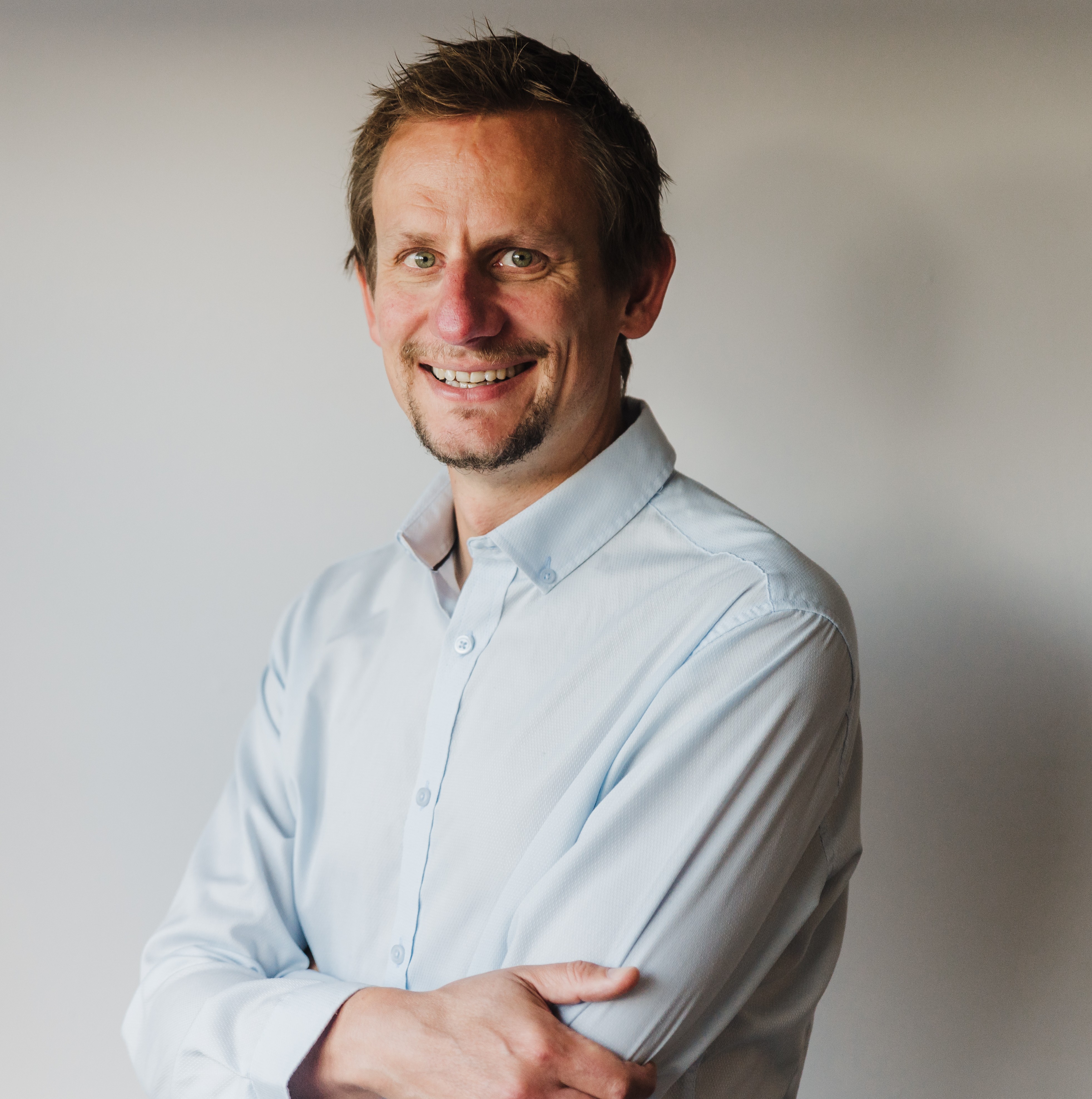 A portrait of Duncan Rusby - A smiling man in a light blue shirt standing with arms crossed against a neutral background.