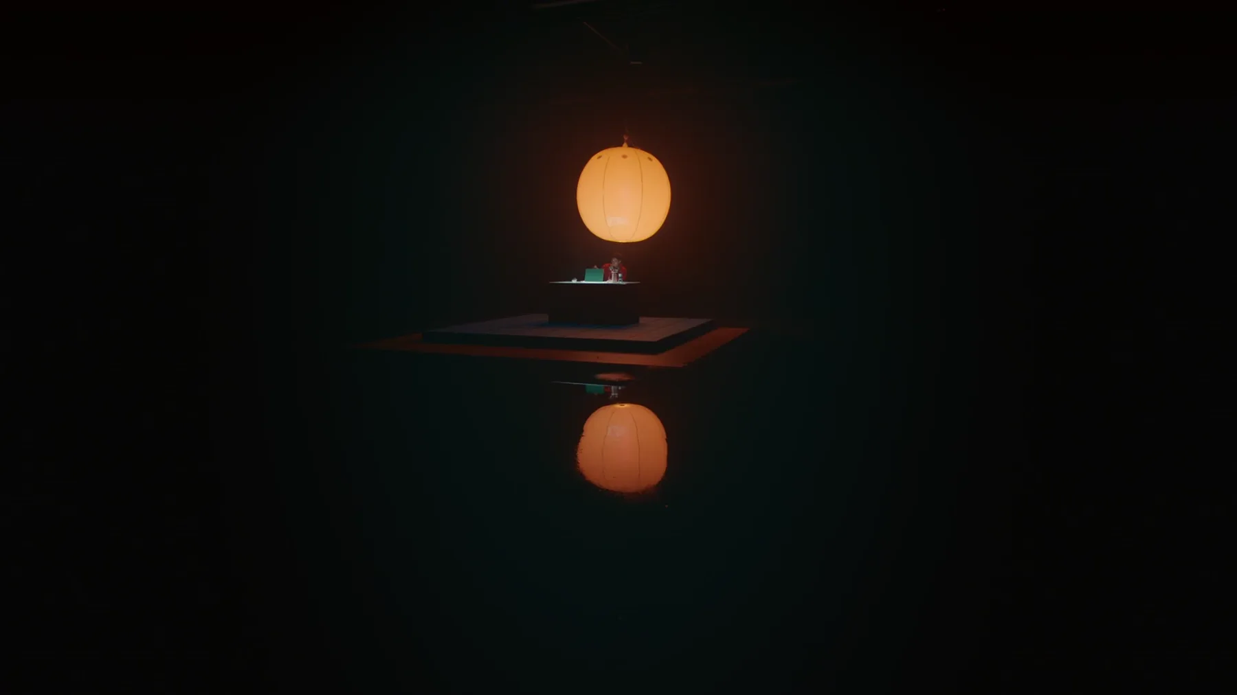 Wide shot of a dark room with an orange pendant lamp hanging above a small platform, a seated figure lit warm below, reflected in a mirrored floor.
