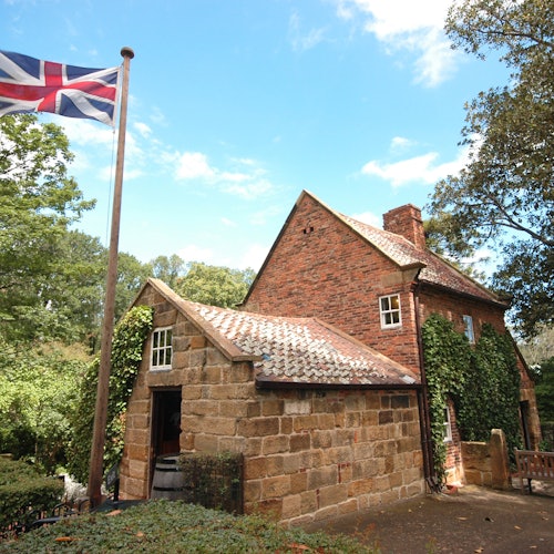 Historic brick house with ivy-covered walls, a stone extension, and a Union Jack flag in the foreground. Trees surround the building.