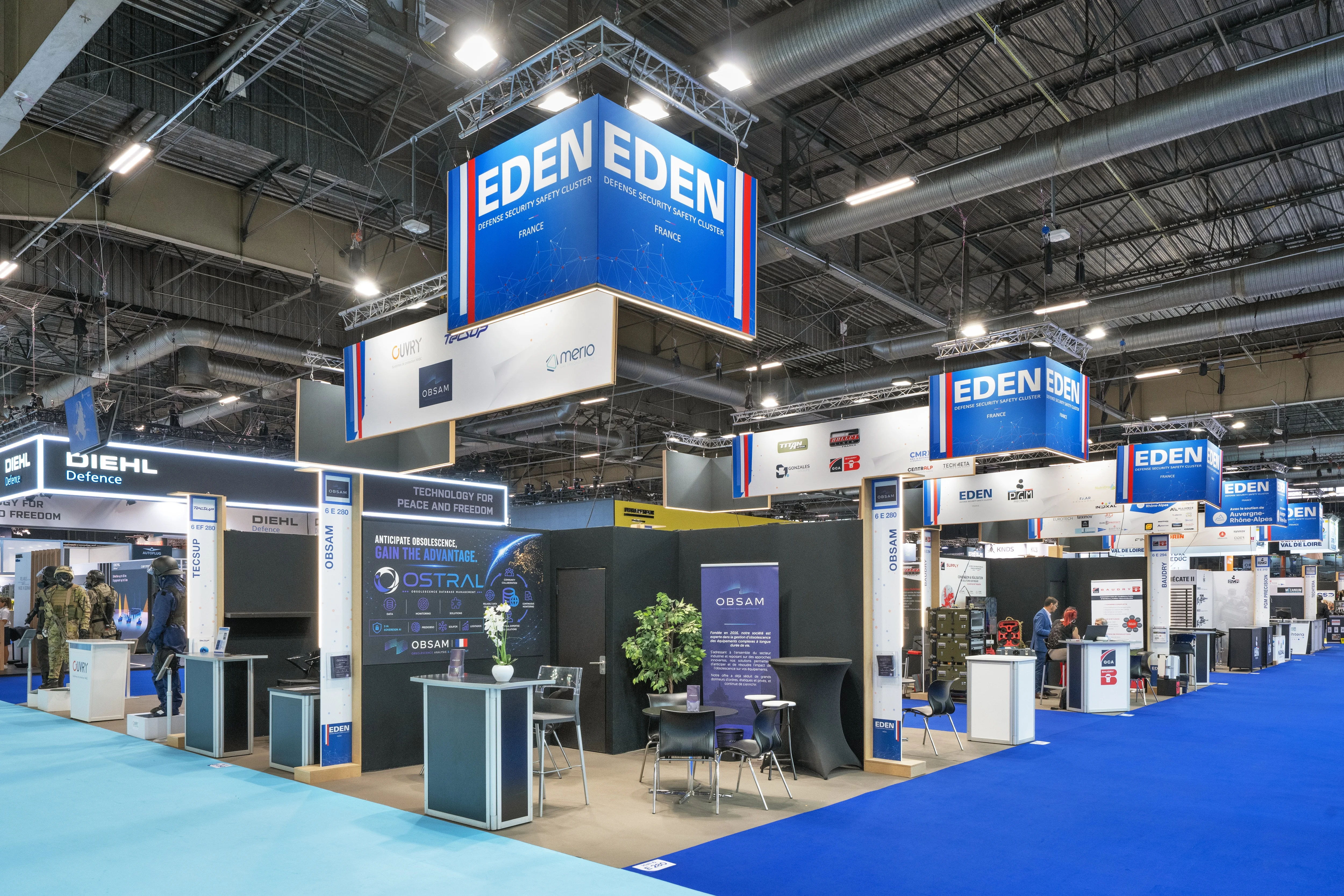 Stand EDEN Cluster conçu et réalisé par Expace à Eurosatory 2026 – pavillon collectif défense bleu-blanc-rouge regroupant les adhérents (SAIB, Sysnav, OBSAM, PGM…) avec vitrines produits, démonstrateurs et accueil central.