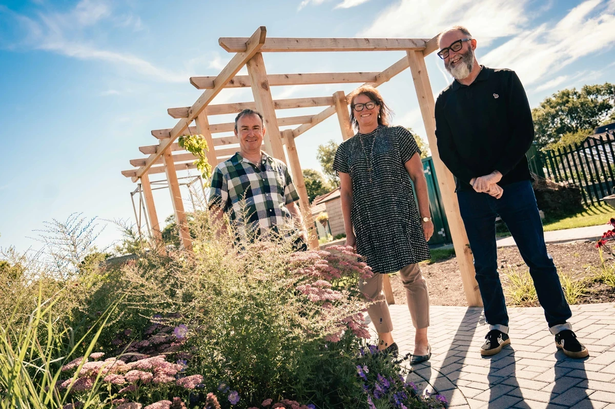 Three people stand outdoors in a landscaped garden area with flowering plants, beneath a wooden pergola structure on a bright sunny day.