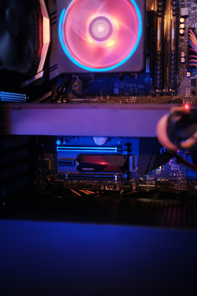 A close-up of a gaming PC's interior showcasing an illuminated cooling fan, a Samsung SSD, and the X470 Gaming Pro Carbon motherboard, highlighting high-performance computer hardware and RGB lighting in a modern gaming setup.