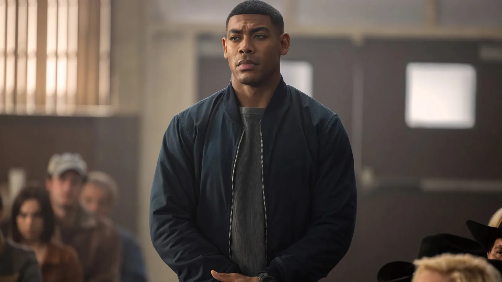 A focused man in a dark jacket stands attentively in a crowded room, surrounded by seated individuals, suggesting a public event or gathering.