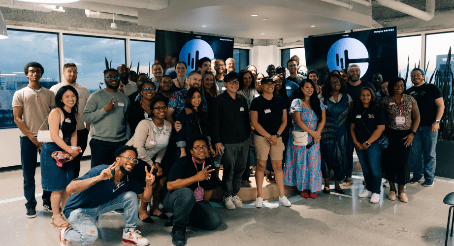 A diverse group of people gathers inside a modern office space with large windows, posing for a group photo in front of two digital screens displaying a stylized logo.