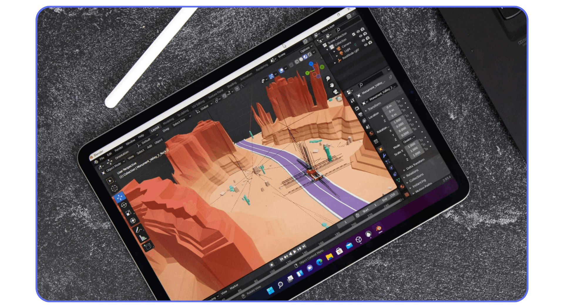 iPad displaying Blender with a low-poly 3D canyon scene, featuring red rock formations and a winding road. The Apple Pencil lies beside the device on a textured gray surface.