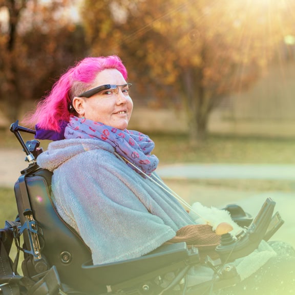 A person with pink hair and glasses smiles warmly in a wheelchair. They're outdoors with autumn trees in the background and sunlight streaming in.