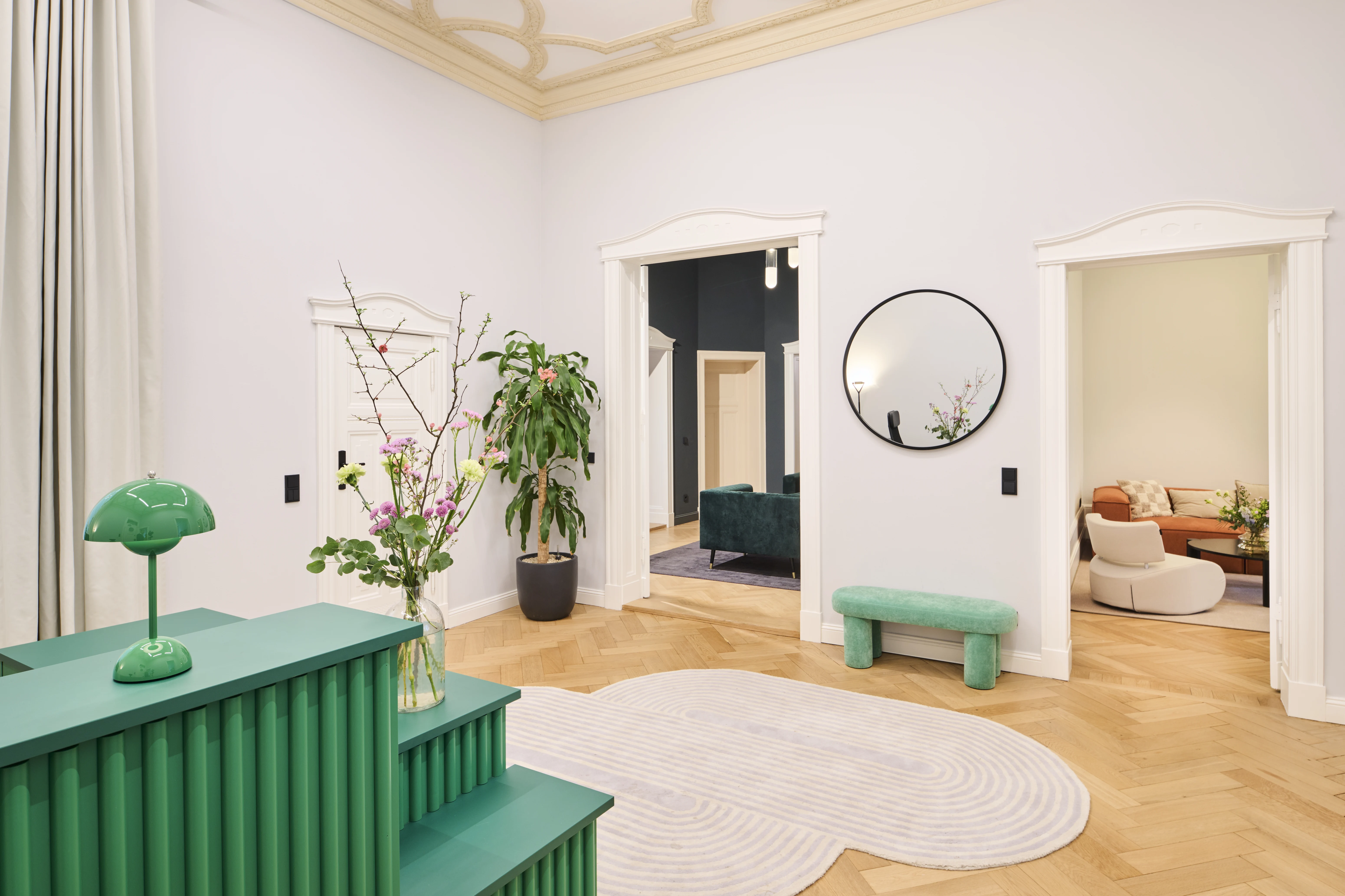 Entrance hall at INTERMATE BERLIN with a green slatted reception counter and matching mushroom lamp, a turquoise velvet bench, round mirror and views into lounge rooms.