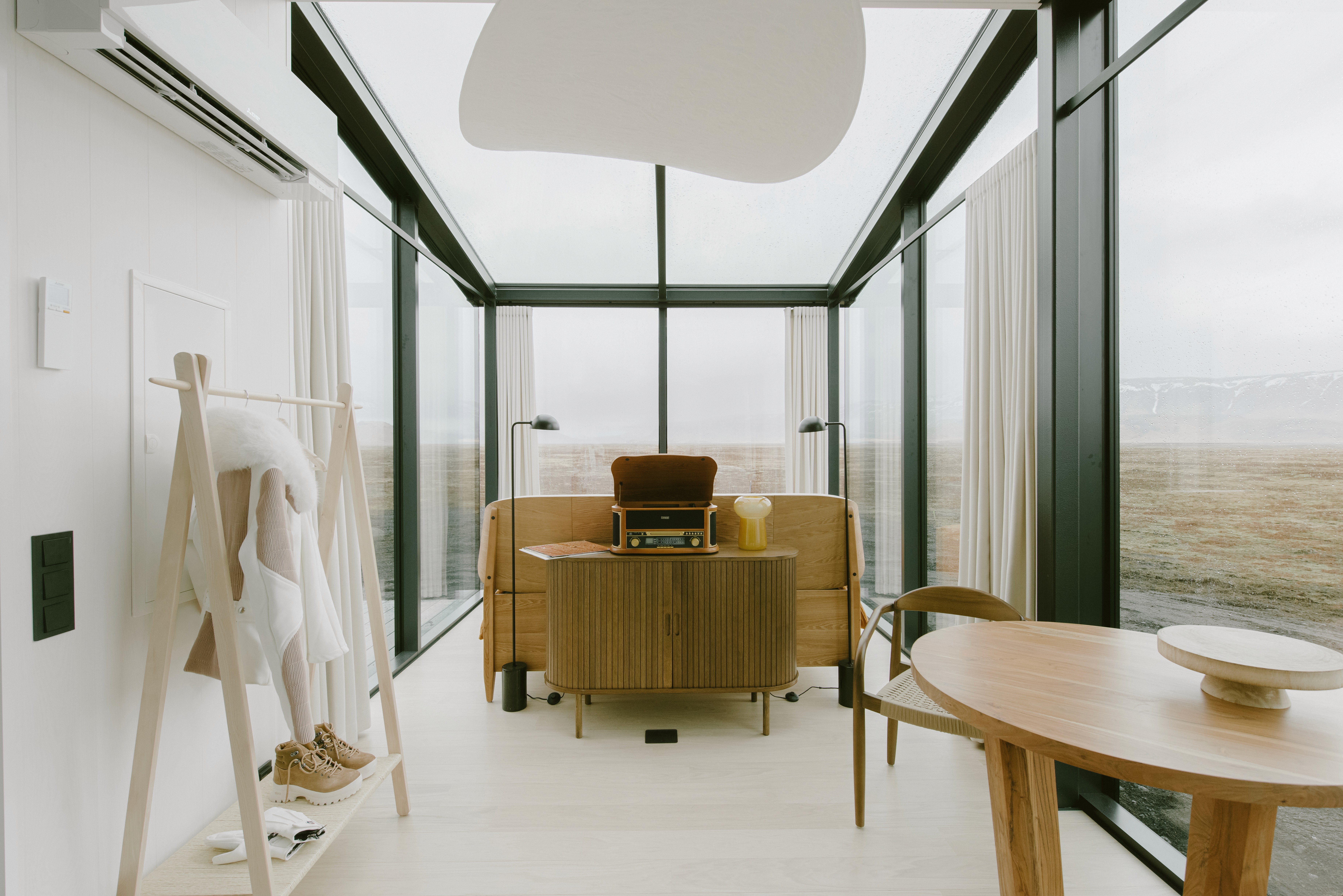 Bright, minimalist interior with floor-to-ceiling glass walls, featuring a wooden bed, compact cabinet with record player, round dining table, and soft furnishings, overlooking a wide, open landscape.