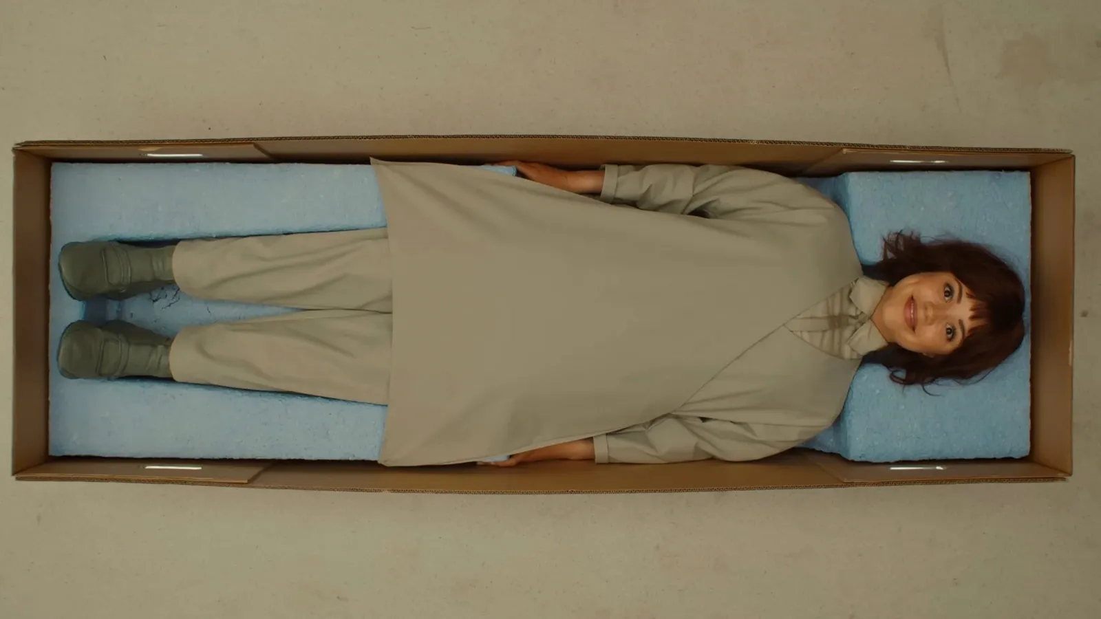 A female woman humanoid lies in a cardboard box, dressed in a beige outfit with green socks, on a light blue cushioned lining, inside an enclosed, minimalist setting.
