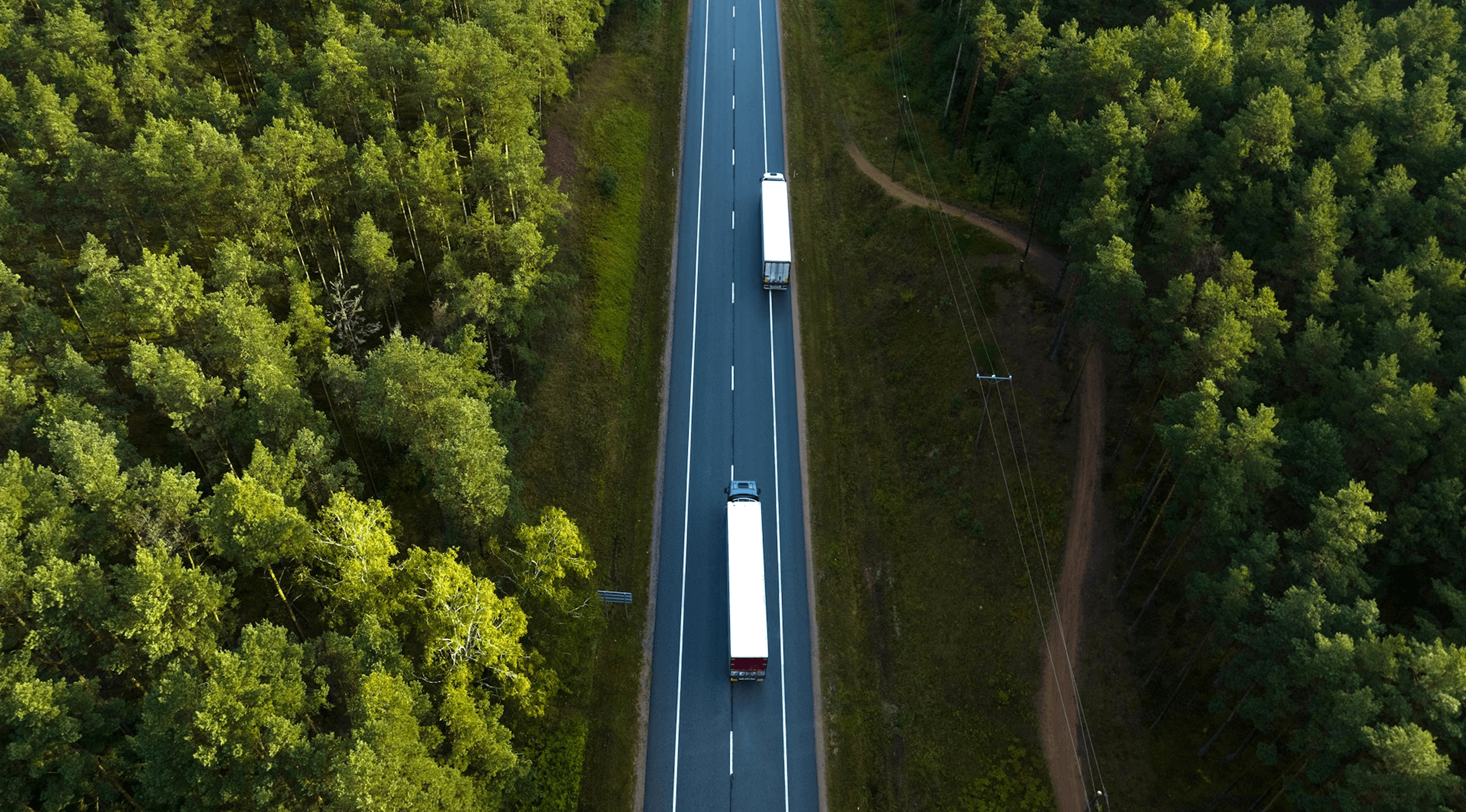 Aerial view of a two-lane highway cutting through dense green forest. Three semi-trucks with white trailers travel in the same direction on the road, spaced out along the highway. Power lines run parallel to the road on the right side. The surrounding forest is lush with deciduous trees in full foliage.