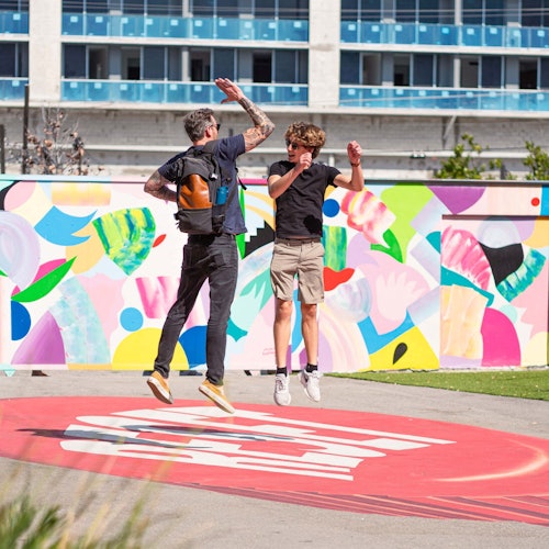 两个人在室外靠近色彩斑斓的壁画跳跃；一个背着背包，另一个穿着短裤。背景中可以看到一座建筑物。