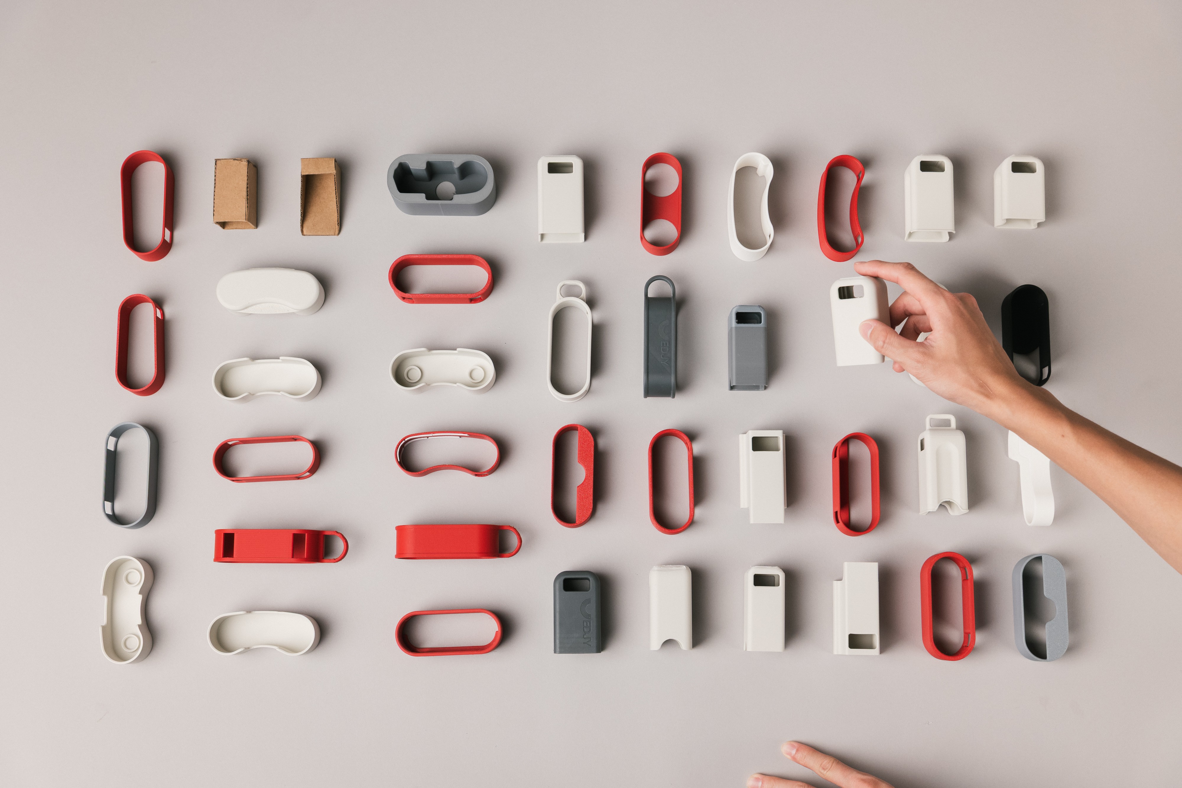 Dozens of EDJY prototype parts laid out in a grid in red, white, gray and black, with a hand reaching in to lift one out.