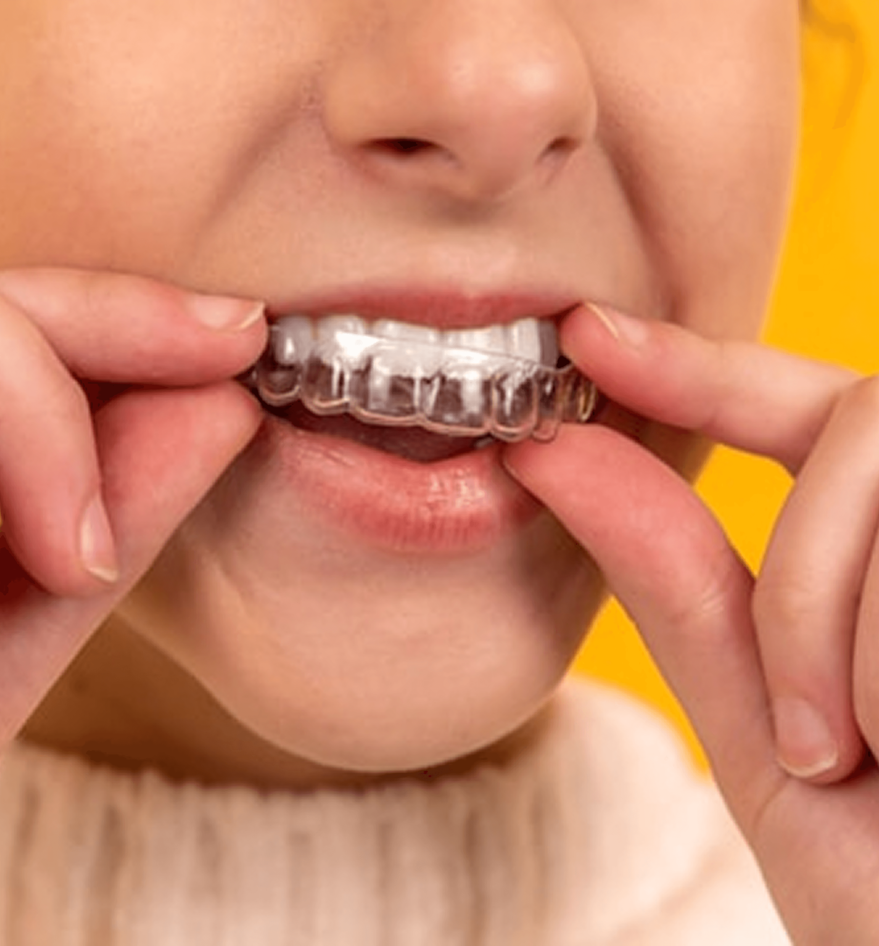 Person inserting clear dental aligner onto upper teeth against yellow background.