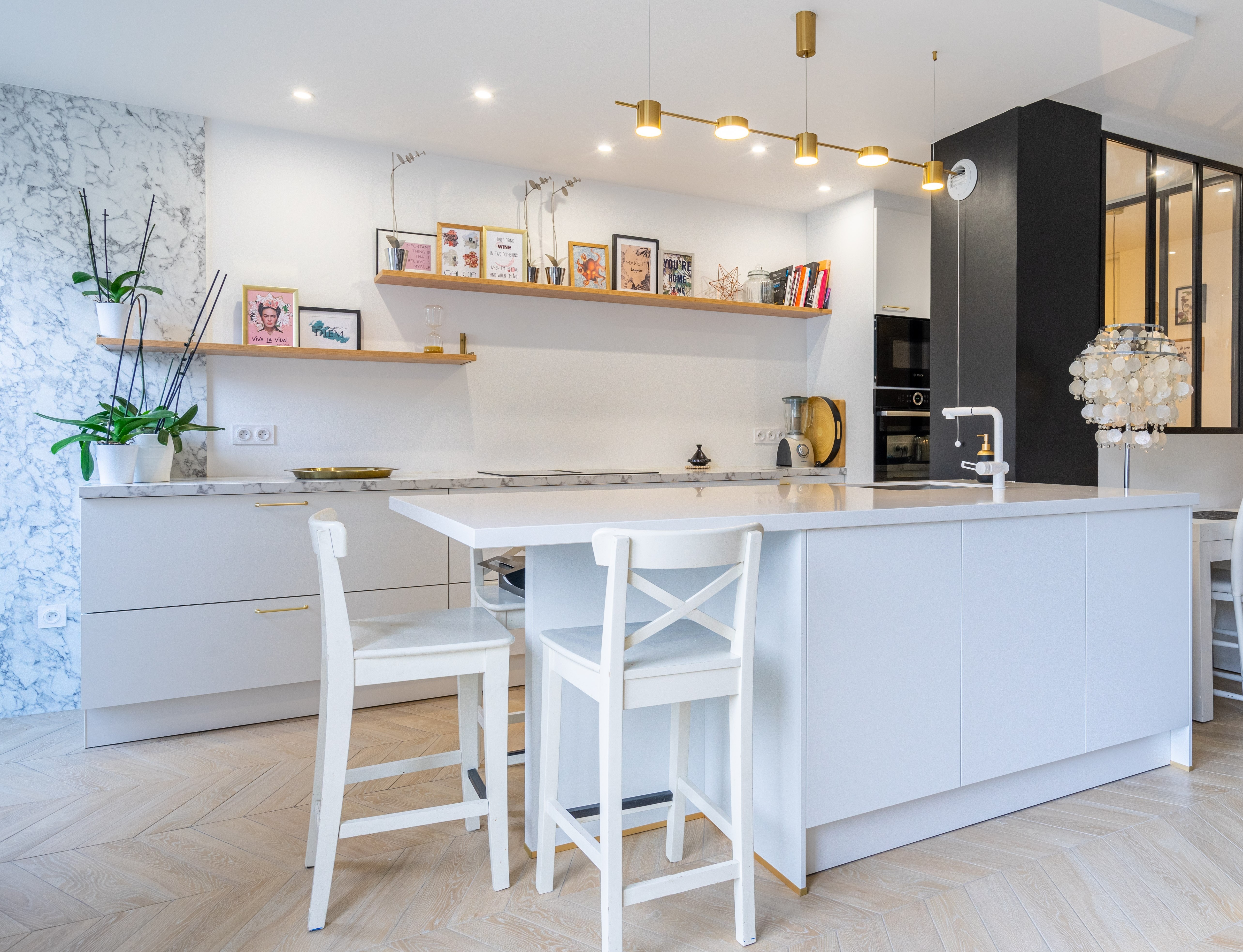 Rénovation cuisine Seine-et-Marne avec armoires blanches, grand îlot, sol bois chevrons et luminaires dorés, mur accent marbre, architecte d'intérieur.