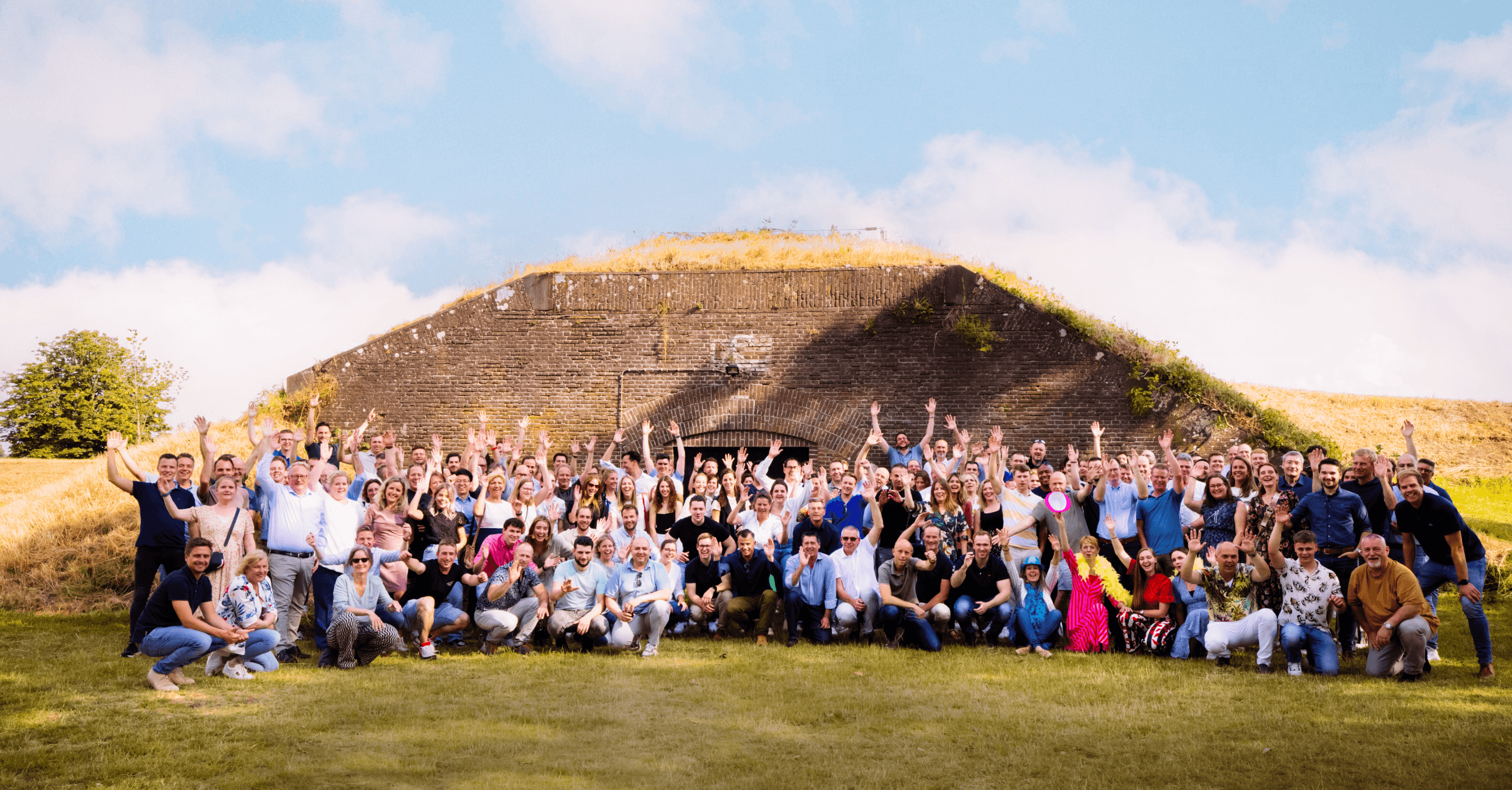 Groepsfoto van het Kader Qnect team voor een historisch fort, met meer dan 100 medewerkers die enthousiast hun handen in de lucht steken. De afbeelding toont de sterke teamgeest en samenwerking binnen Kader Qnect, een bedrijf dat zich richt op innovatieve oplossingen. De groene omgeving en blauwe lucht dragen bij aan de positieve sfeer van dit teamuitje. Perfect voor het benadrukken van de bedrijfscultuur en teamactiviteiten van Kader Qnect.