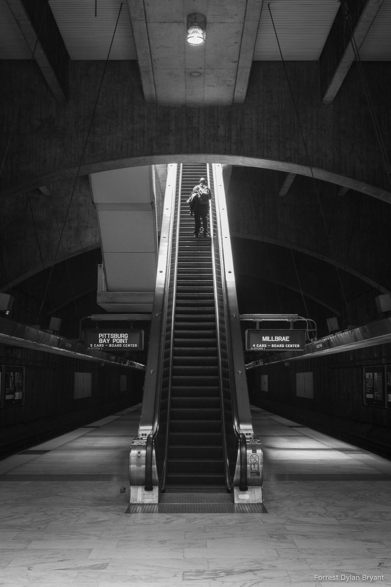 A lonely subway platform. On an escalator, a man carrying heavy bags rises toward the light.