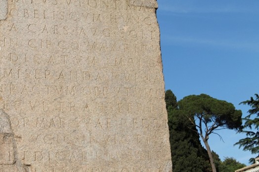 An old stone wall with engraved letters, placed near a green park with trees.