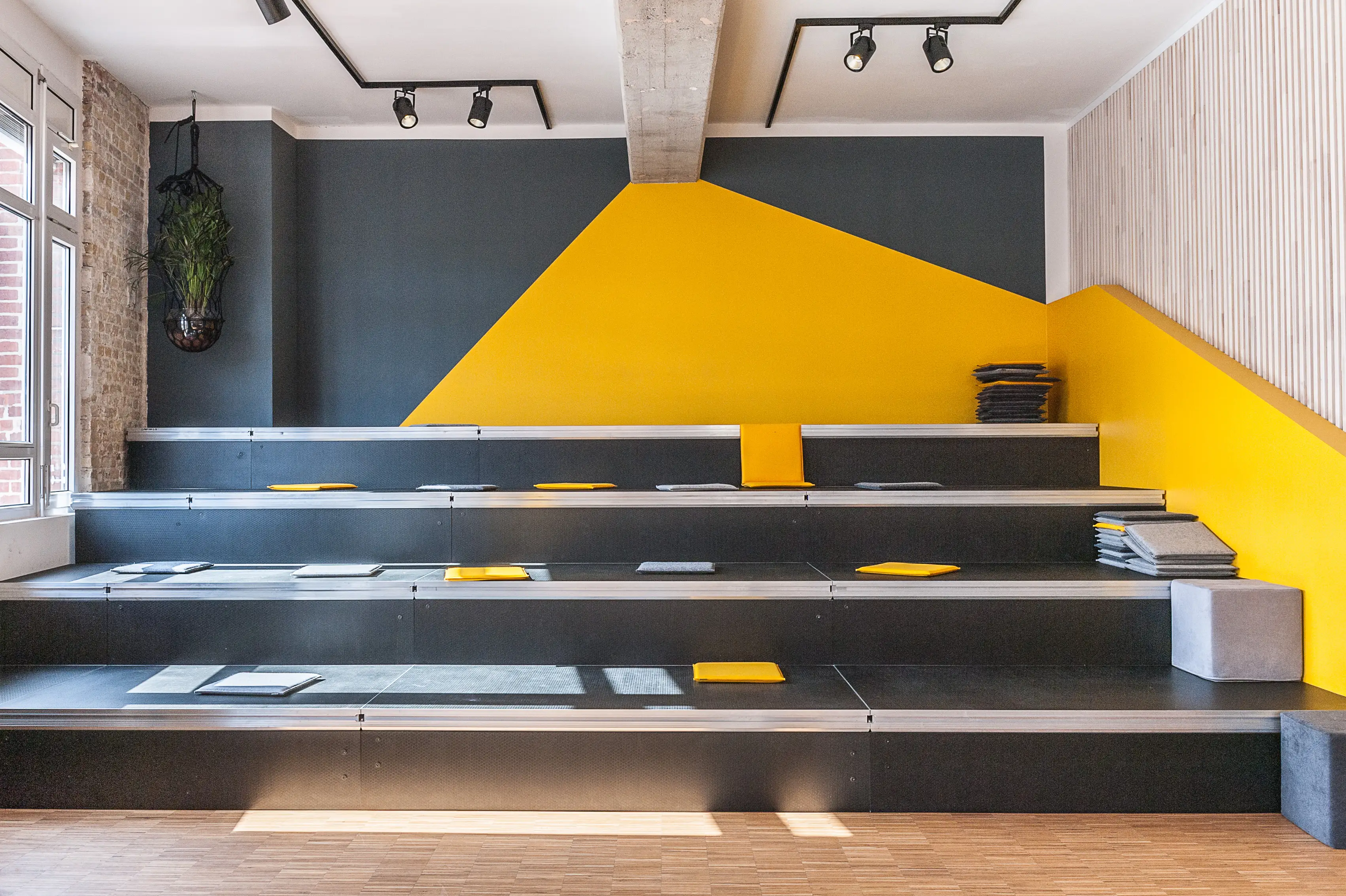 Stepped seating tribune at BASILICOM in dark grey with yellow and grey felt cushions, a graphic yellow wall section, black spotlights on the ceiling and a hanging plant by the window.