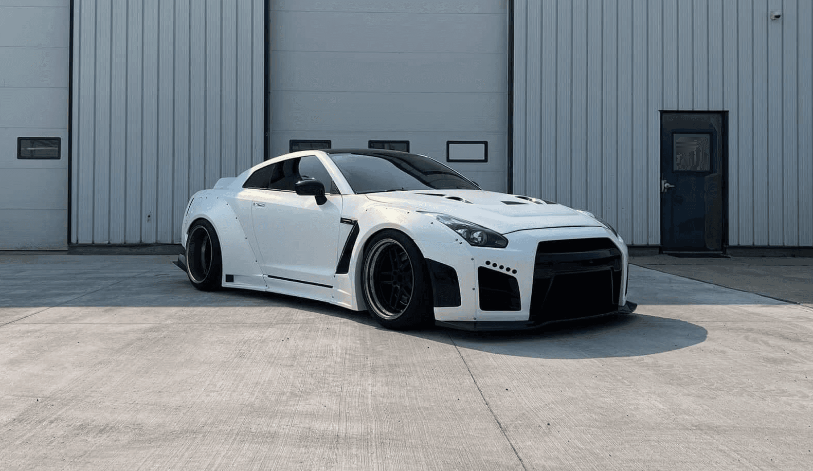 A white Nissan GT-R with a widebody kit is parked on a concrete driveway in front of a white industrial building.